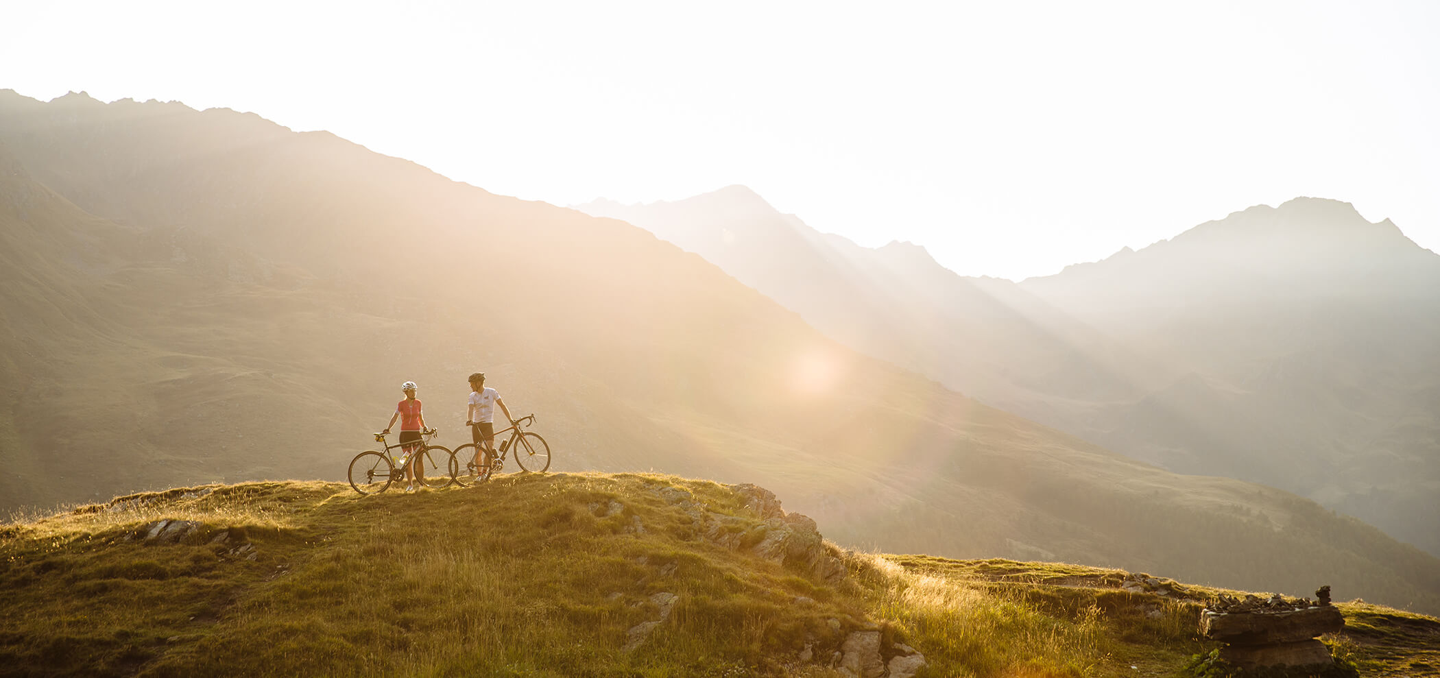 Zwei Personen mit Fahrrädern stehen auf einem grasbewachsenen Bergrücken, umgeben von einer weiten Berglandschaft im goldenen Sonnenlicht.