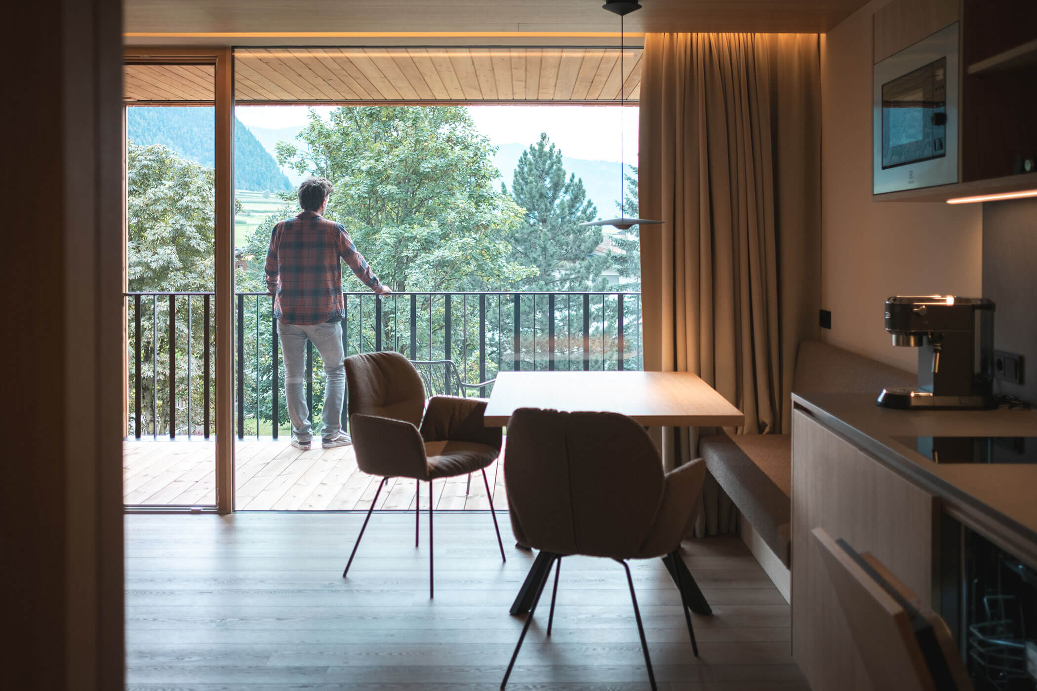 Kitchenette and dining table in the foreground, in the background a man standing on the balcony enjoying the view - Gisse Zwölf